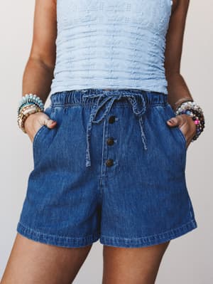 A close mid-shot of denim pull-on shorts with a relaxed fit, paired with a powder blue textured crop top and stacked beaded bracelets — casual and collected with a warm, lived-in feel.