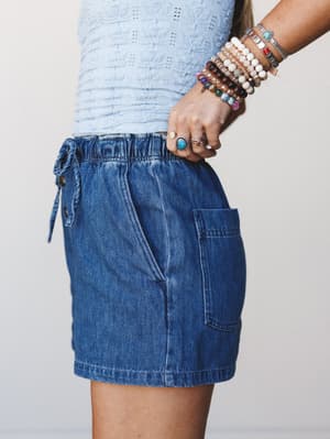 A close-cropped side view of a denim mini skirt with a drawstring waist and patch pocket, paired with a light blue textured top and a stacked arm full of beaded and stone bracelets — casual and collected, with just enough personality in the accessori