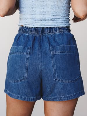 A back-view close-up of relaxed denim shorts with large patch pockets and an elastic waistband, paired with a light blue textured smocked top — easy, lived-in summer dressing.