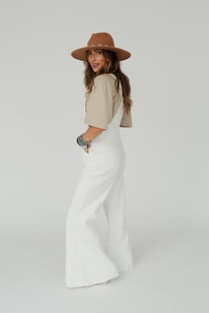 A woman in a beige shirt and the Milly Distressed Hem Wide Leg Overall in cream smiles over her shoulder, wearing a wide-brimmed brown hat and several bracelets, set against a plain white background.