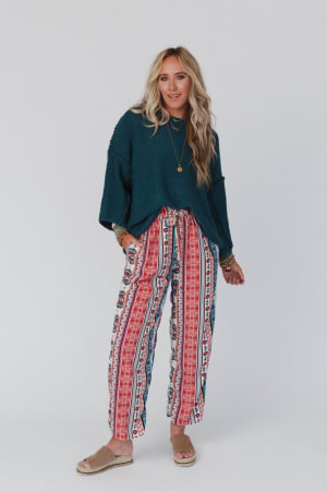 A woman with long blonde hair wears The Nest Staycation Wide Leg Drawstring Pant in Red Clay, a teal oversized sweater, tan sandals, and gold necklaces. She stands against a white background, smiling with one hand in her pocket.