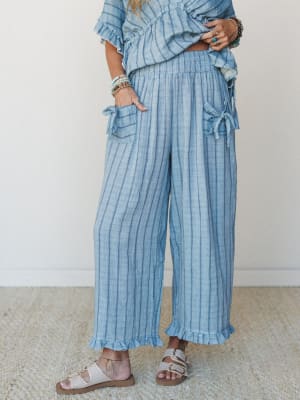Wide-leg striped linen-look pants in a soft denim blue, styled as a matching set with a ruffled top, stacked turquoise bracelets, and flat sandals — the kind of outfit that looks equally at home on a boardwalk or a back patio.