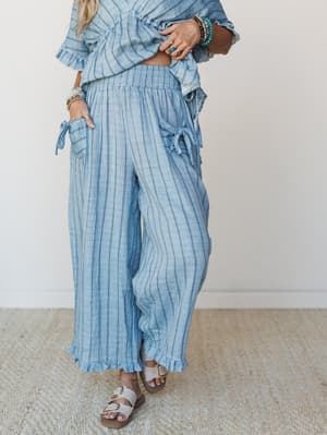 A breezy striped wide-leg pant in washed denim blue styled as a matching set with a coordinating ruffle top, layered boho jewelry, and sandals — the kind of outfit that looks equally at home on a coastal patio or a farmers market morning.