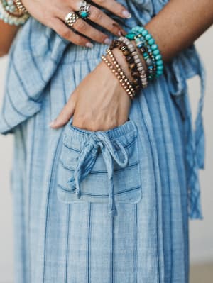 A close-up of a woman's hand tucked into the patch pocket of soft chambray striped pants, layered with a stack of boho bead bracelets in turquoise, tiger's eye, and gold — the kind of detail shot that makes you reach for your jewelry box.