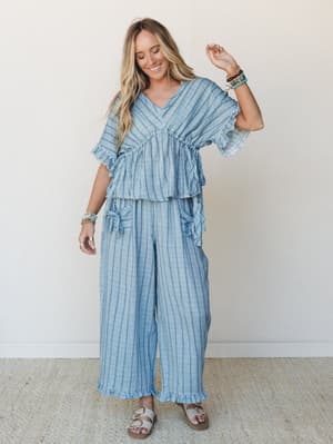 A matching striped linen-look set in soft denim blue — the wide-leg pants and relaxed ruffle-hem top move together like they were made for a slow beach morning or a long lunch with good people.