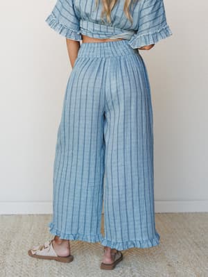 A back-view shot of wide-leg striped pants in a soft denim blue, styled as a matching set with a cropped wrap top — the ruffle hem and smocked waist give it that breezy, effortless feel that lives somewhere between a beach vacation and a really good