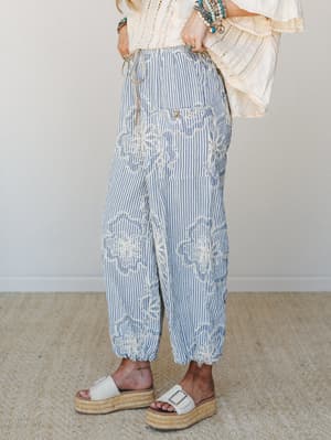 A close crop of relaxed, balloon-silhouette striped pants in denim blue and cream, covered in looping floral embroidery — styled with a lace bell-sleeve top, stacked turquoise bracelets, and white espadrille platform sandals for an effortlessly boho