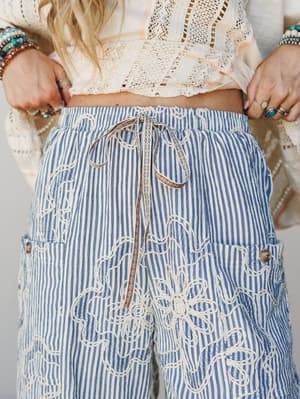 A close-up detail shot of wide-leg striped pants in denim blue and cream, covered in chunky floral embroidery and finished with a woven drawstring tie — styled with a crochet crop top and stacked boho jewelry for a lived-in, expressive look.