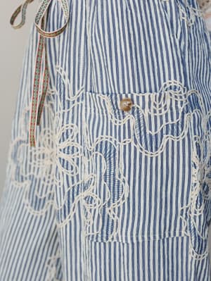 A close-up detail shot of blue and white striped pants with loopy white embroidery cascading down the leg and a button-front patch pocket — the kind of piece that makes people stop you mid-errand to ask where you got them.