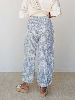 A back-view shot of wide-leg striped pants with cream floral embroidery, styled with a lace-trimmed cream top and platform espadrilles — easy and expressive, like a summer morning with nowhere to be.