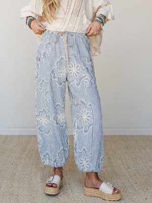Wide-leg striped pants in denim blue with cream floral embroidery take center stage, styled with a crochet lace top, stacked turquoise bracelets, and espadrille platform sandals for a breezy, well-traveled feel.