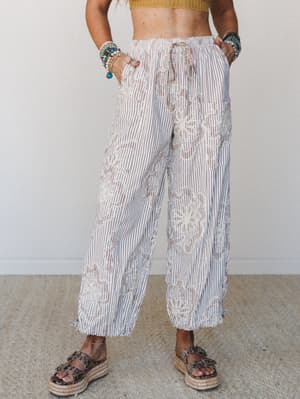 Wide-leg striped pants with all-over floral embroidery styled with a cropped mustard knit top, stacked beaded bracelets, and platform espadrille sandals — relaxed and expressive with a distinctly boho-summer feel.