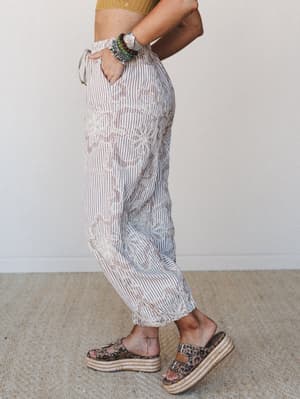 A side-profile shot of relaxed, tapered pants in a cream and mocha stripe with tonal swirl embroidery, styled with a cropped mustard knit top, stacked boho bracelets, and leopard print platform espadrilles — easy and expressive all at once.