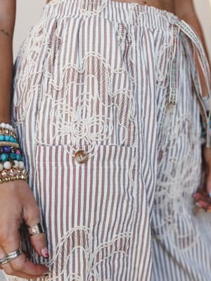 A close-up detail shot of wide-leg striped pants in mocha and cream, featuring intricate cream embroidery and a front patch pocket with a single button closure — styled with stacked beaded bracelets and silver rings that feel collected, not coordinat