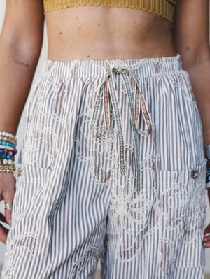 A close-up detail shot of wide-leg striped pants with intricate crochet embroidery and a patterned drawstring tie, styled with a mustard crop top and stacked bracelets — the kind of pants you'd buy for a trip and then wear everywhere.