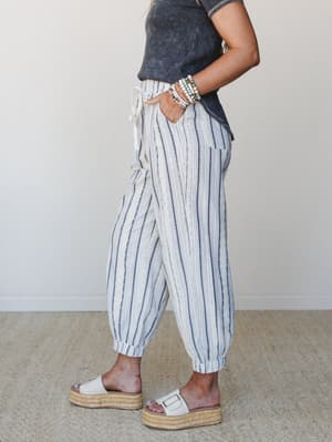 A side-profile shot of relaxed striped jogger pants in cream and navy, styled with a washed charcoal tee and white platform espadrille sandals — the kind of outfit that works equally hard at the farmers market and on a beach boardwalk.