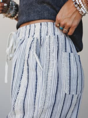 A close-up detail shot of textured striped pants with mixed lace and woven panels, styled with a cropped navy tee and layered gemstone jewelry — relaxed and well-traveled in the best way.