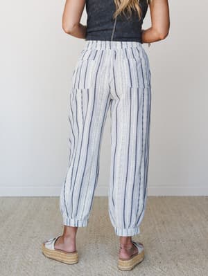 Shot from behind, a model shows off relaxed striped jogger pants in a cream and navy woven stripe — paired with a dark ribbed tank and espadrille platform sandals, the whole look reads effortlessly easy, like a beach town Saturday with nowhere to be.