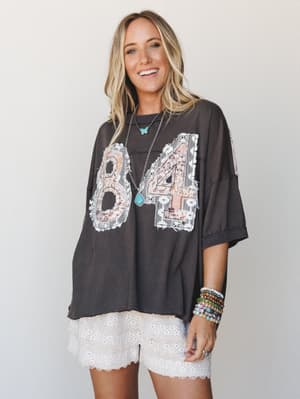 An oversized washed-charcoal graphic tee with lace and floral appliqué numbering paired with white eyelet shorts — the kind of effortless, personality-packed outfit she'd throw on and somehow look completely intentional.