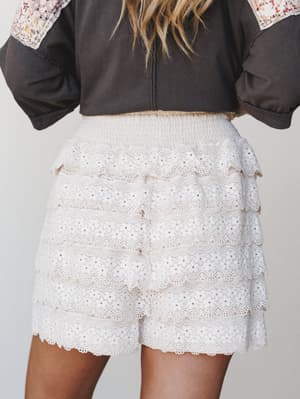 A back-view close-up of tiered white eyelet lace shorts with a smocked waistband, styled under a charcoal patchwork top — romantic and effortlessly boho with serious summer energy.