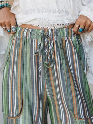 A close-up of multicolor striped wide-leg pants with a relaxed drawstring waist, styled with a white lace crop top and layered silver and turquoise rings — easy, expressive, and very much vacation-ready.