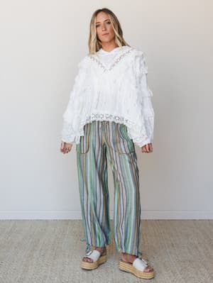A white ruffled lace blouse paired with relaxed multicolor striped pants in earthy greens and neutrals — the kind of outfit that feels equally at home at a farmers market or a rooftop dinner.