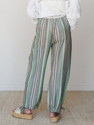 Shot from behind, a model wears relaxed wide-leg striped pants in earthy greens, slate, and amber paired with a ruffled white lace top and platform espadrilles — the kind of outfit that works just as well at a seaside market as it does on your back p