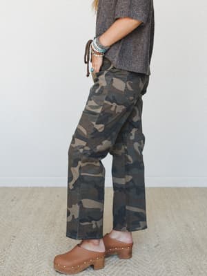 A side-profile shot of wide-leg camo pants styled with a chunky brown sweater, wood-sole clogs, and a stack of beaded bracelets — the kind of effortless, earthy outfit that looks like it came together without trying.