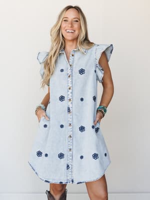 A light-wash denim shirt dress covered in navy floral embroidery, styled with stacked beaded bracelets and cowboy boots — the kind of dress that makes a Tuesday feel like something worth getting dressed for.