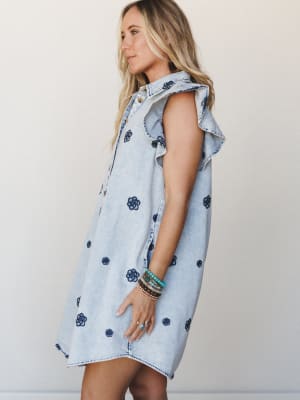 A light-wash denim mini dress with all-over dark navy floral embroidery and flutter sleeves, styled with stacked bracelets — the kind of dress that makes a farmer's market feel like an occasion.