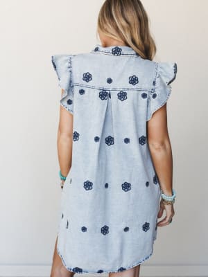 A light wash denim shirt dress covered in dark navy floral embroidery, shot from the back to show off the ruffle sleeves and center pleat — styled with stacked beaded bracelets that feel effortlessly collected.