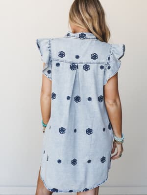 A light wash denim shirt dress covered in dark navy floral embroidery, shot from the back to show off the ruffle sleeves and center pleat — styled with stacked beaded bracelets that feel effortlessly collected.
