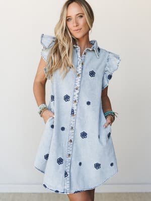 A light-wash denim shirt dress with scattered navy floral embroidery and ruffle cap sleeves — the kind of dress that works as hard at a weekend farmers market as it does at a summer birthday dinner.