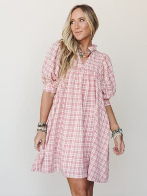 A breezy pink plaid mini dress with puff sleeves and a ruffle neckline, styled with layered beaded jewelry for a playful, warm-weather feel.