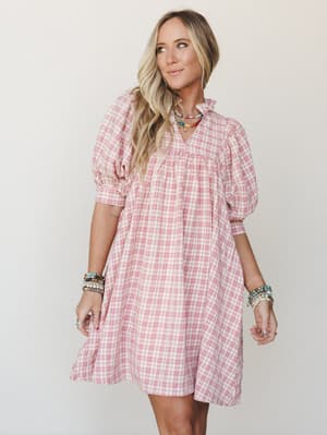 A breezy pink plaid mini dress with puff sleeves and a ruffle neckline, styled with layered beaded jewelry for a playful, warm-weather feel.