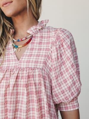 A close-up of a pink plaid babydoll-style dress in a seersucker-like texture, styled with layered colorful beaded necklaces — sweet and a little nostalgic, like a picnic in the best possible way.