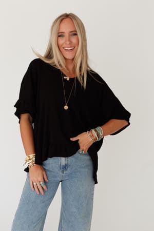 A woman with long blonde hair smiles at the camera, wearing The Nest Let Me Be Ruffled Top - Black with light blue jeans and colorful accessories, standing against a plain white background.