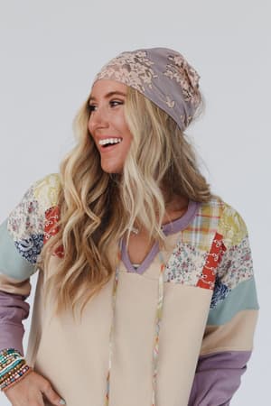 A smiling woman with long, wavy blonde hair wears the Sarah Floral Lace Headscarf in gray and a colorful patchwork top. She looks to the side, showing off several beaded bracelets on her wrist against a plain white background.