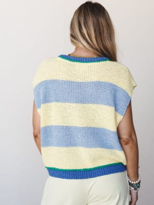 Shot from the back, a model wears a chunky knit stripe sweater vest in soft butter yellow and periwinkle blue with bright green contrast trim at the neckline and hem, paired with cream trousers — easy, colorful, and effortlessly warm-weather-ready.
