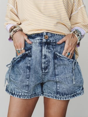 A close mid-shot styled around acid wash denim shorts with oversized drawstring cargo pockets, paired with a yellow striped sweatshirt and stacked with layers of boho rings and beaded bracelets — the kind of outfit that feels effortless but clearly i
