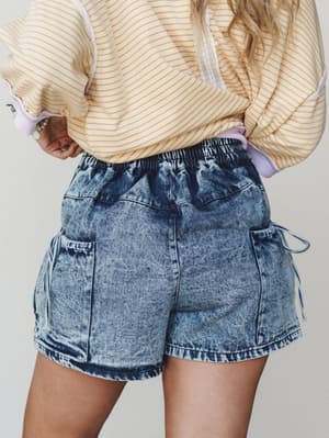 A close mid-shot from behind showing acid wash denim cargo shorts with an elastic waistband, styled with a yellow striped oversized sweatshirt — casual and a little nostalgic, like a weekend that has no plans.