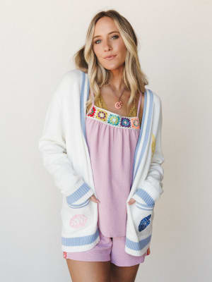 A cream knit cardigan with periwinkle stripe trim and embroidered floral pocket details layered over a lavender cami top with colorful granny square crochet detailing — playful, soft, and full of personality.
