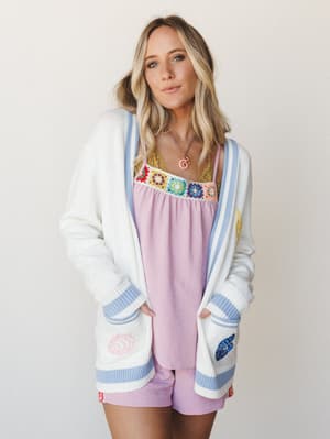 A cream knit cardigan with periwinkle stripe trim and embroidered floral pocket details layered over a lavender cami top with colorful granny square crochet detailing — playful, soft, and full of personality.