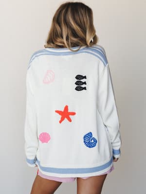 A breezy, back-facing shot of an oversized white knit cardigan with playful ocean-themed embroidery — seashells, starfish, and tiny fish — trimmed in periwinkle stripe, paired with lavender shorts for a summery, carefree feel.
