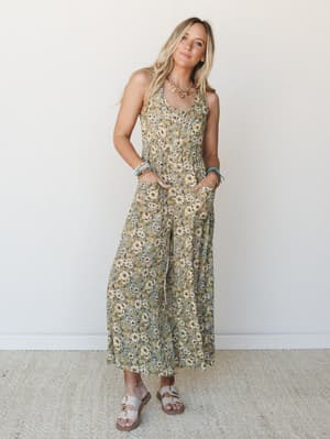 A flowy wide-leg floral jumpsuit in olive and golden yellow worn with layered boho jewelry and buckle sandals — the kind of outfit that looks equally at home at a farmers market or a rooftop dinner.