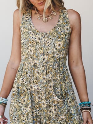 A close-cropped shot of a floral olive jumpsuit with button-front detail, styled with stacked turquoise bracelets and a gold coin necklace — the kind of outfit that looks like it was made for a slow Saturday with nowhere to be.