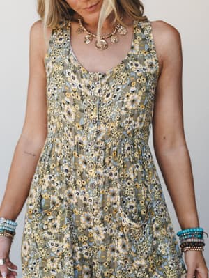 A close-cropped shot of a floral olive jumpsuit with button-front detail, styled with stacked turquoise bracelets and a gold coin necklace — the kind of outfit that looks like it was made for a slow Saturday with nowhere to be.