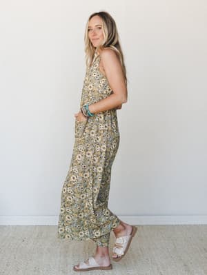 A flowy floral jumpsuit in an olive-toned daisy print styled with white buckle sandals and stacked beaded bracelets — the kind of outfit that looks equally at home on a farmers market morning or a summer dinner patio.