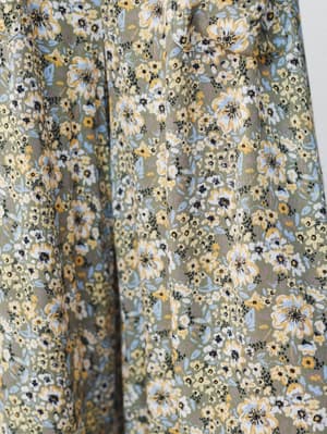 A close-up of a flowy floral fabric in a soft olive base, covered in a dense garden of yellow, white, and periwinkle blooms — the kind of print that feels like wandering through a wildflower field in late summer.