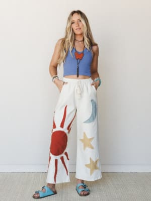 A breezy, expressive outfit pairing a periwinkle ribbed crop tank with wide-leg cream pants featuring bold sun, moon, and star appliqués — the kind of look that feels like it was made for someone who takes their personal style seriously and their cof